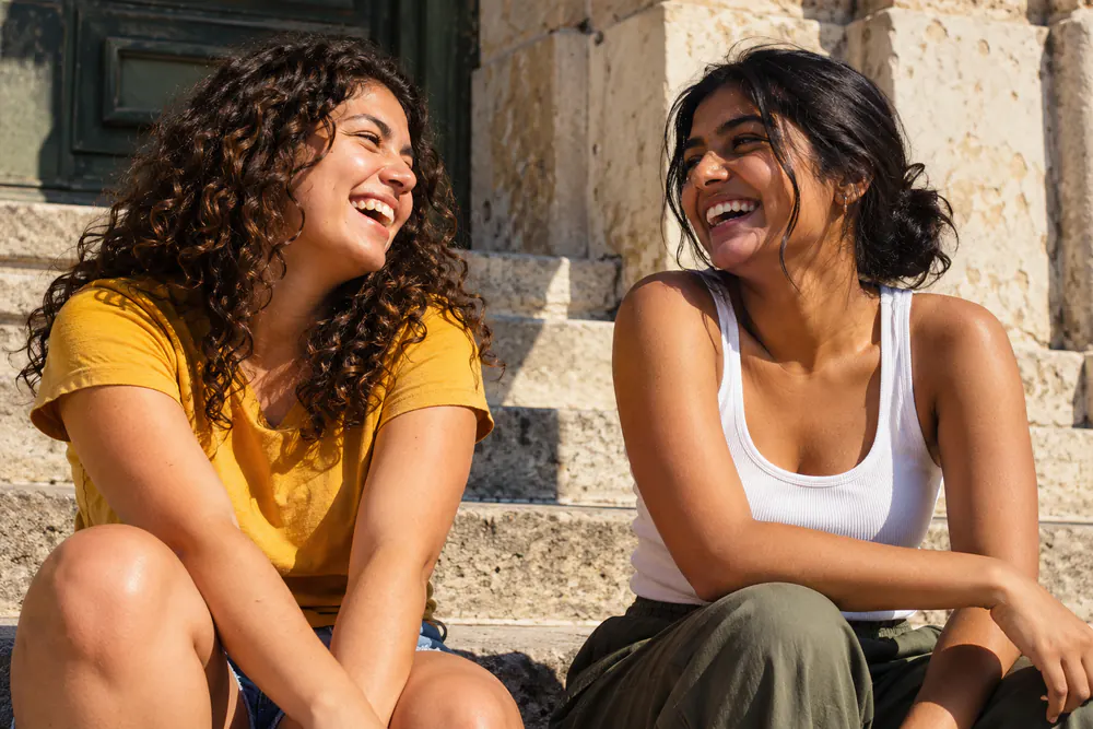 Two friends laughing on a sunlit stone step.