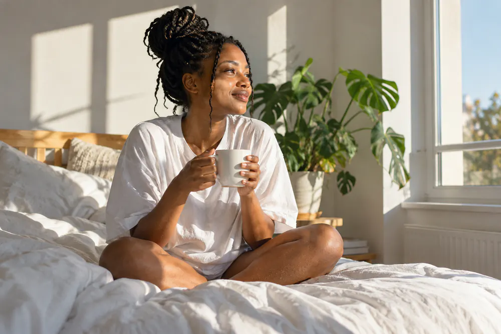 A woman with braids sits cross-legged in a bright bed with a mug, looking out of the window.