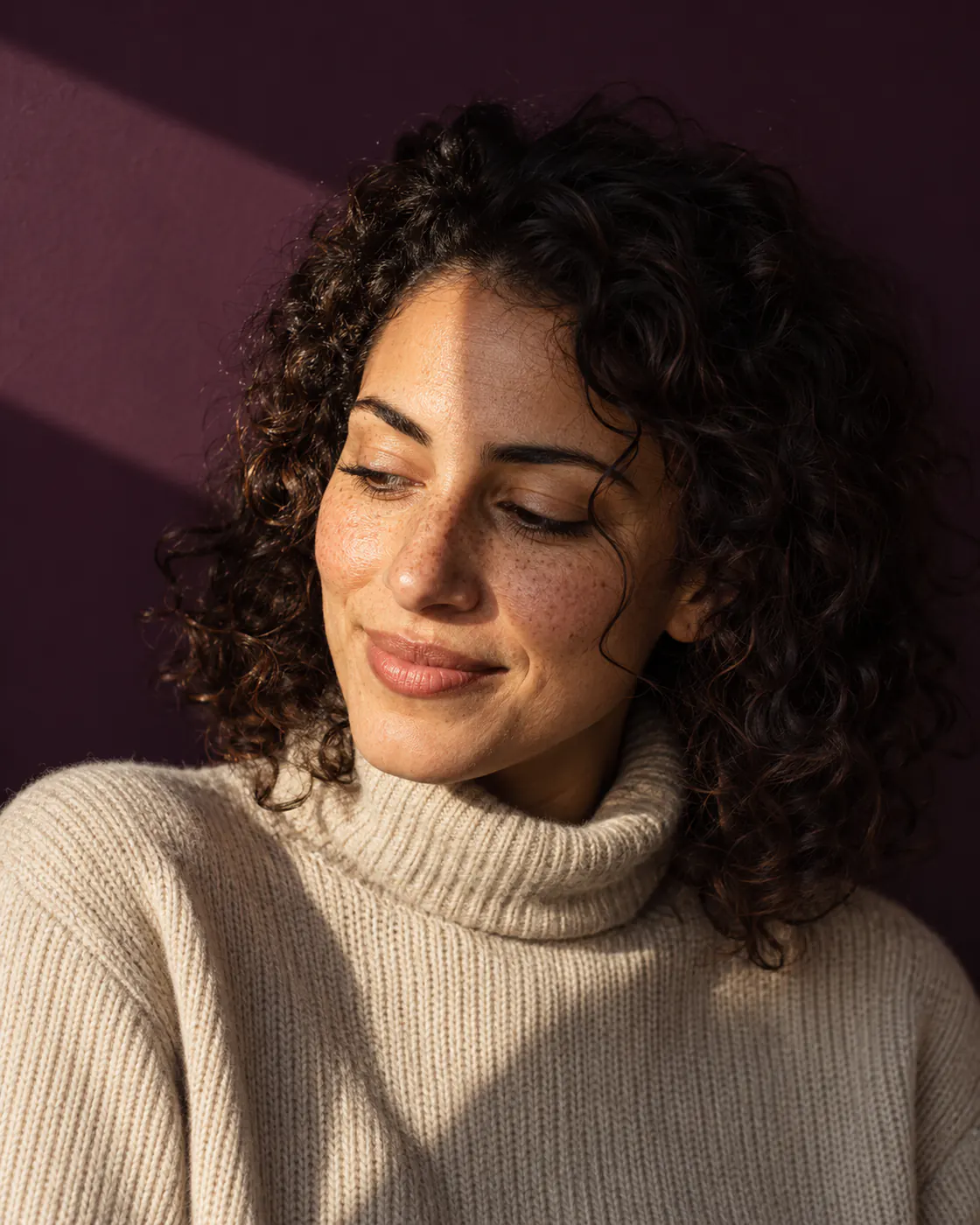 A woman with curly hair and freckles smiles to herself in a slice of warm light against dark aubergine paper.