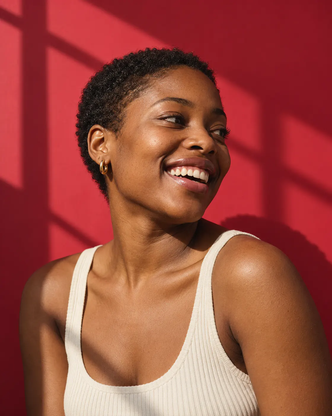 A young woman with short natural hair laughs, looking away, against cherry-red paper in hard window light.
