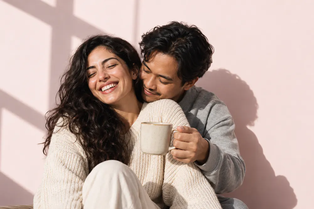A man rests his chin on a laughing woman's shoulder and holds out a mug to her, against pale blush paper.
