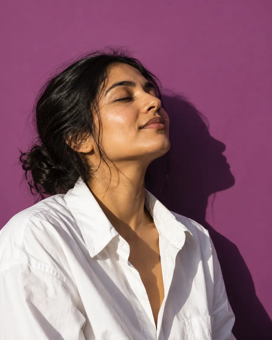 A woman in a white shirt with her eyes closed, face tilted up into the sun, against plum paper.