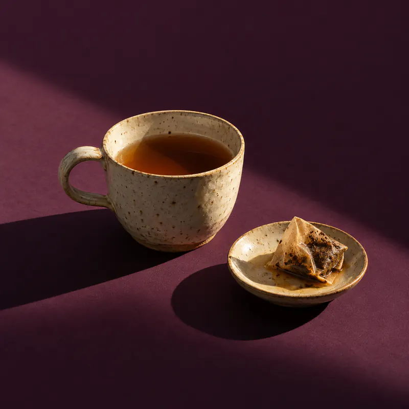 A speckled ceramic cup of tea gone cold in a beam of evening light on dark aubergine paper.